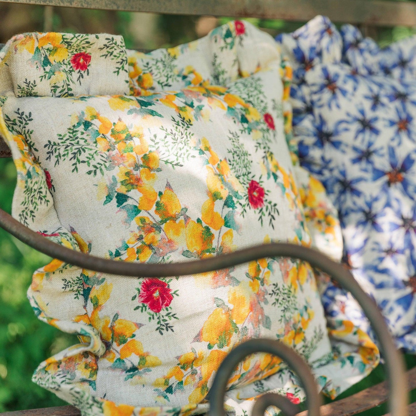 A square pillow with ruffles made of organic linen and printed with bright florals sits on an outdoor bench, the sun playing on its surface.